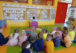 dzieci siedzą na dywanie w dwóch rzędach jedna dziecko siedzi tyłem do nich na pufce